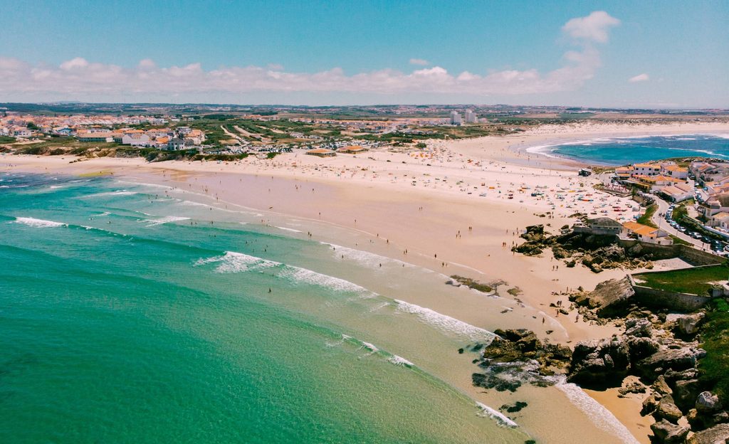 A wide, sandy north-facing beach curving around a low headland, with small waves breaking close to shore