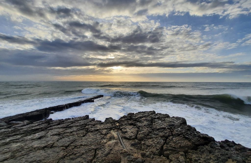 Waves breaking over a rocky point at sunset, with clouds over the Atlantic