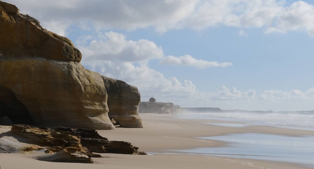 A wide, calm sandy beach with dunes and a distant golf-resort backdrop