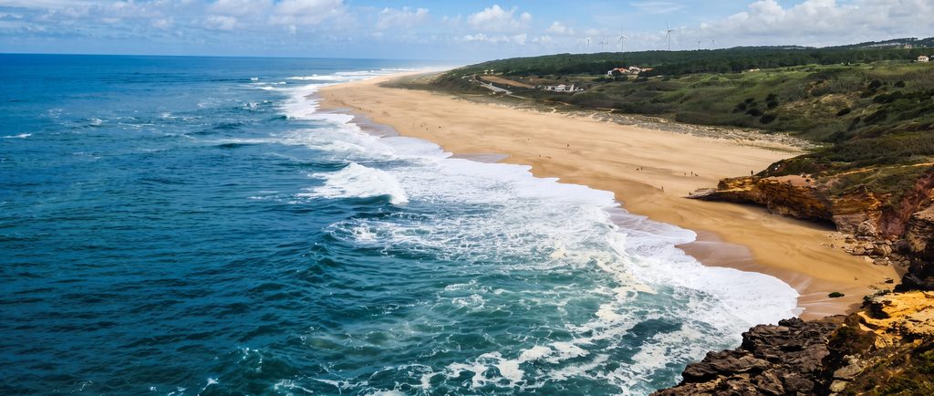 A sandy beach beneath eroded cliffs near Nazaré, with waves rolling in