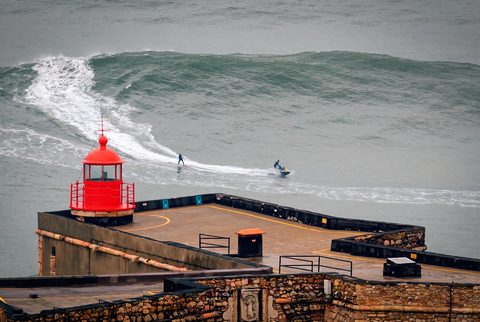 A tow-in surfer and jet-ski support rider passing the red lighthouse at the Nazaré fort