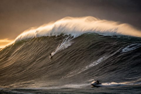 A big-wave surfer carving down a huge wave in front of the red lighthouse, watched by a crowd of spectators